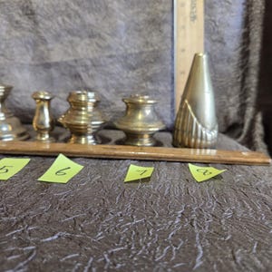 May include: A collection of antique brass objects, including small candle holders and a decorative vase. A wooden ruler is placed next to the objects for scale. The objects are arranged on a textured brown surface.