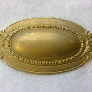 May include: A gold-toned oval-shaped decorative drawer pull with ornate detailing around the edges.