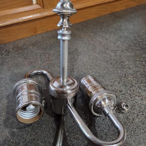 May include: A silver-toned lamp fixture with two bulb sockets and a decorative finial. The lamp has a curved arm design and a central support column. The sockets are designed to hold standard light bulbs.