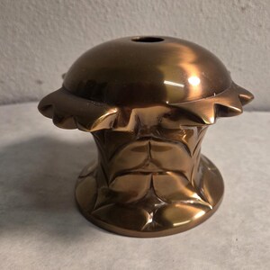 May include: A bronze-colored, decorative object with a rounded top and a flared base. The top has a small hole and a scalloped edge. The base features a repeating leaf pattern. The object has a metallic sheen and appears to be made of metal.
