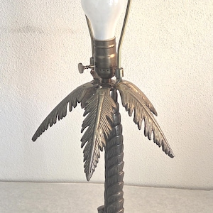 May include: A bronze-colored palm tree lamp with a white bulb. The lamp has a detailed palm tree trunk and fronds, and a round base. The lamp is plugged in with a black cord.