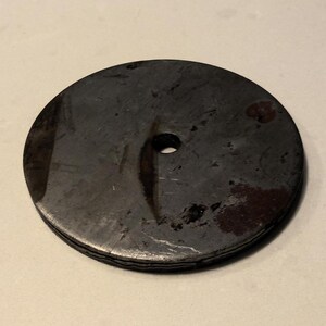 May include: A round, flat, metal disc with a small hole in the center. The disc is dark gray with a slightly textured surface.