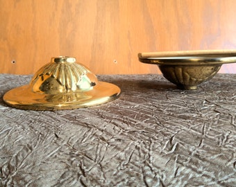 May include: Two antique brass objects sit on a textured surface. One is a hat-shaped piece with a wide brim and a central hole. The other is a bowl-shaped object with a floral design.