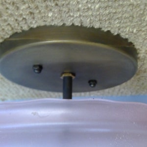 May include: A ceiling light fixture with a frosted pink glass shade and a dark metal base. The shade has a wavy design. The fixture is mounted on a textured, light-colored ceiling.