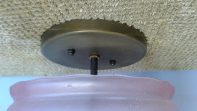 May include: A ceiling light fixture with a frosted pink glass shade and a dark metal base. The shade has a wavy design. The fixture is mounted on a textured, light-colored ceiling.