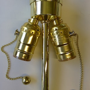 May include: A gold-toned three-way lamp socket with a chain pull switch. The socket has a decorative finial on top and two side sockets. The chain is attached to a small ball.