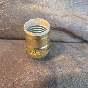 May include: A gold-colored lamp socket with a paper-lined interior and a silver-colored threaded base. The socket has a cylindrical shape with a ridged design around the base. The item is likely for a light fixture.