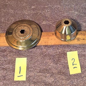 May include: Two brass objects with hexagonal openings are displayed. One is a flat, circular piece with concentric rings, and the other is a conical shape. A wooden ruler with inch markings is also present. Yellow sticky notes labeled "1" and "2" are below the objects.