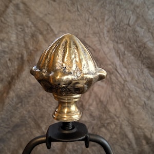 May include: A gold-coloured lamp finial. The finial has a textured, cone-shaped top with a decorative base and a black metal screw. The background is a textured, brown fabric.