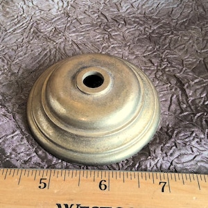 May include: A gold-toned metal lamp finial with a round base and a center hole. The finial has a decorative, raised rim.