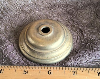 May include: A gold-toned metal lamp finial with a round base and a center hole. The finial has a decorative, raised rim.