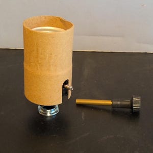 May include: A brown cardboard lamp socket with a silver base and a small metal screw. A separate gold-coloured metal rod with a black knob is next to the socket. The items are on a dark surface.