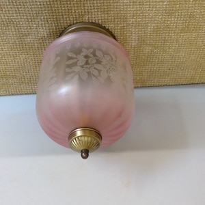May include: A vintage pink glass light fixture with a floral design etched on the surface. The fixture has a brass-colored top and bottom, and a ribbed pattern on the lower portion. The light fixture is designed to be mounted on a wall or ceiling.