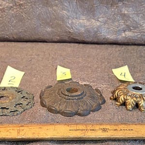 May include: Five vintage metal lamp parts are displayed. The parts include a silver, conical shade, a decorative, circular piece, a dark brown, floral-shaped piece, a gold-colored, leaf-shaped piece, and a silver, flat disc. Each part has a central hole.