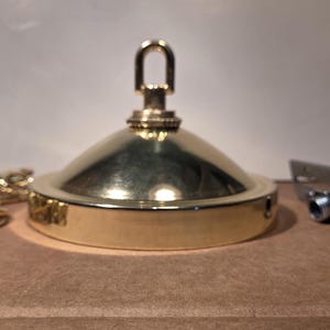 May include: A polished brass ceiling canopy kit featuring a dome-shaped cover with a central loop for suspension. The set includes a matching gold-toned metal chain and a silver-colored mounting bracket with a threaded pipe for installation.