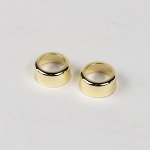 May include: Two gold-colored metal rings, each with a smooth, rounded surface. The rings are similar in size and shape, and they are positioned side-by-side on a white background.