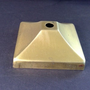 May include: A gold metal square base with a round hole in the center. The base is slightly raised in the center, creating a pyramid shape.