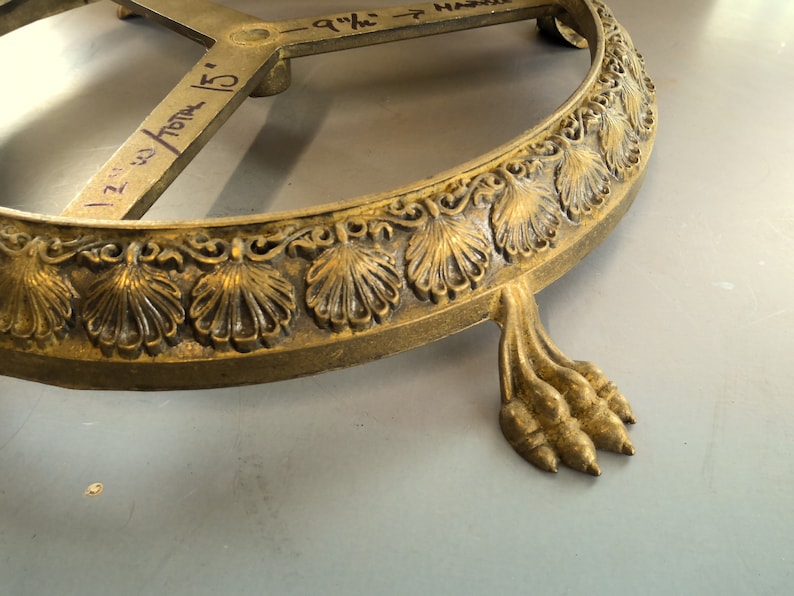 Puede incluir: Una base de metal dorado con un borde festoneado decorativo y tres patas de garra. La base tiene un di&aacute;metro de 23 cm y tiene una inscripci&oacute;n manuscrita en la parte superior.