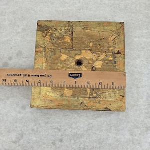 May include: A square, wooden furniture leg with a distressed gold finish. A ruler is placed across the top, measuring approximately 3 inches wide. The leg has a hole drilled through the center.