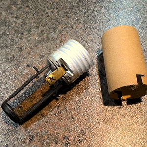May include: A disassembled light fixture component set against a mottled background. The set includes a silver-colored bulb socket with a brass screw and a black metal frame, along with a brown cardboard sleeve.