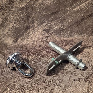May include: A silver-toned metal mounting bracket featuring a threaded central post and a flat, slotted crossbar, accompanied by a separate, matching circular cap with a small, integrated loop for attachment purposes.