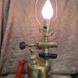 May include: A unique lamp crafted from a vintage brass blowtorch. The lamp features a red handle, a black valve, and a decorative finial. The light bulb is enclosed in a metal frame, creating a warm, inviting glow. The lamp is a blend of industrial design and functional art.
