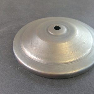 May include: A brushed metal ceiling canopy with a central hole. The round canopy has concentric circular ridges and a matte finish. The object is silver and sits on a gray surface.