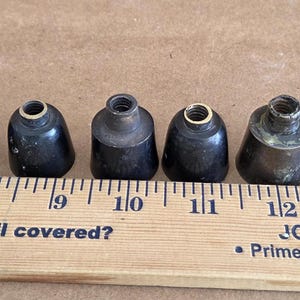 May include: Six antique metal lamp parts, varying in colour from black to brass, are arranged in a row. A wooden ruler with inch markings and the text "JOB PREP" is below the parts. The parts are approximately 2.5 cm in height.