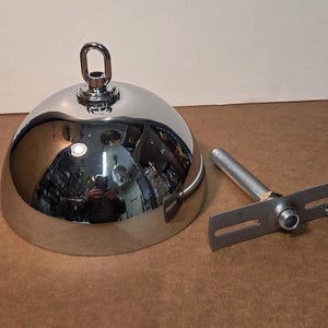 May include: A polished chrome pendant light fitting with a domed shade and a hanging loop. Includes a mounting bracket with a threaded rod and a small green screw. The reflective surface mirrors the surroundings.