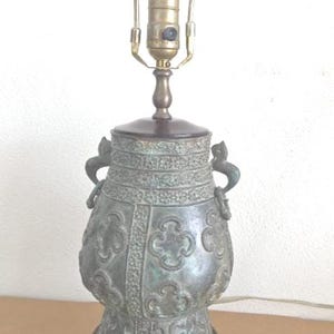 May include: An antique bronze-colored table lamp with a decorative urn-shaped base. The lamp features ornate detailing, including floral patterns and handles. The lamp has a dark brown wooden base and a brass lamp holder. The lamp is approximately 20 inches tall.