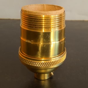 May include: A brass-colored lamp socket with a threaded exterior and a textured base. The top opening is lined with a tan-colored cardboard material. The socket is designed for electrical use.