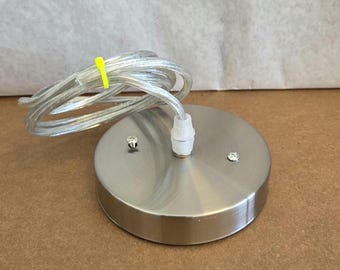 May include: A brushed nickel ceiling canopy with a clear electrical cord. The round canopy has two small screws and a white plastic cord connector. A yellow cable tie secures the cord.