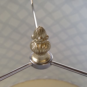 May include: A gold-colored metal lamp finial with a decorative design. The finial is attached to a silver metal lamp base.