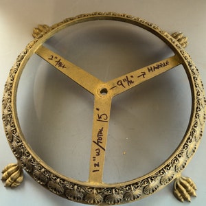 Puede incluir: Un soporte decorativo de metal dorado con una parte superior redonda y tres patas. El soporte tiene un dise&ntilde;o decorativo alrededor del borde y tres brazos con medidas escritas a mano: "2" pulgadas, "1 1/2" pulgadas total / 15", y "9 1/4" pulgadas hasta el m&aacute;rmol".