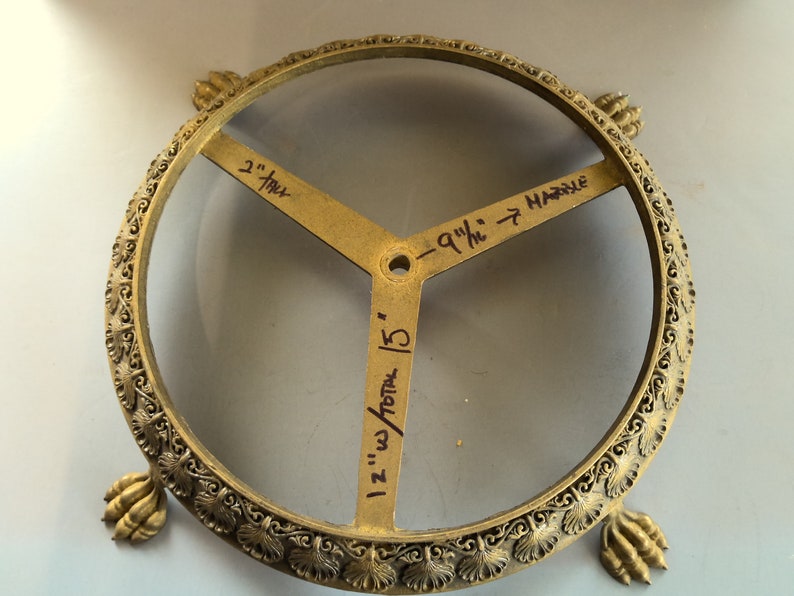 Puede incluir: Un soporte decorativo de metal dorado con una parte superior redonda y tres patas. El soporte tiene un dise&ntilde;o decorativo alrededor del borde y tres brazos con medidas escritas a mano: "2" pulgadas, "1 1/2" pulgadas total / 15", y "9 1/4" pulgadas hasta el m&aacute;rmol".