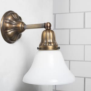 May include: A brass wall sconce with a white glass shade. The sconce has a decorative, fluted design and a bell-shaped shade.