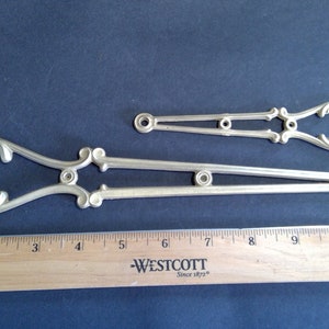 May include: Two ornate metal brackets, one longer than the other, with decorative scrollwork and a circular opening at one end. The brackets are silver in colour and measure approximately 10 inches and 5 inches in length.