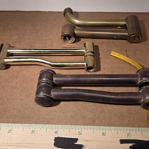 May include: Three metal swing arm lamp components with hinged joints. One is dark brown, one is polished brass, and one is antique brass. A wooden ruler at the bottom shows the arms are approximately 5 to 6 inches in length. A yellow wire is attached.