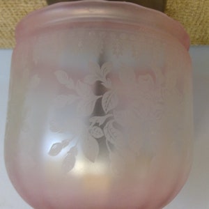 May include: A frosted glass light fixture shade with a soft pink hue. The shade features an etched floral design. The fixture is a cylindrical shape with a slightly flared top. The shade is made of glass.