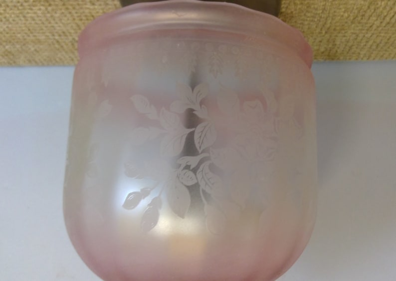 May include: A frosted glass light fixture shade with a soft pink hue. The shade features an etched floral design. The fixture is a cylindrical shape with a slightly flared top. The shade is made of glass.
