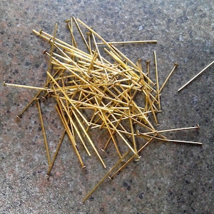 May include: A collection of gold-colored sewing pins scattered on a speckled gray surface. The pins have small, flat heads and long, slender shafts. The pins are used for sewing and crafting.