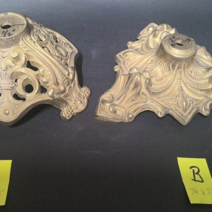 May include: Two gold-toned metal lamp bases with ornate designs. The base on the left is 6 1/2 inches by 3 inches. The base on the right is 7 1/4 inches by 2 1/2 inches.