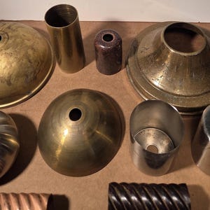 May include: A collection of assorted metal lamp parts including brass and silver-toned domes, cylindrical sleeves, a dark brown connector, and two textured, spiraled metal tubes arranged in a group on a flat surface for lighting repair.