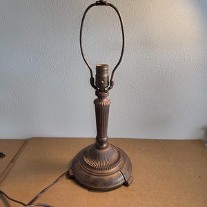 Peut inclure: Une lampe de table de couleur bronze avec une base décorative et un support incurvé pour l'abat-jour. La lampe a une colonne cannelée et une base ronde. Le cordon électrique et la prise sont visibles.