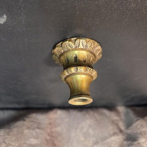 May include: A brass-colored furniture finial with a decorative leaf pattern around the top. The finial has a threaded base and a turned, hourglass-shaped body. The finish has a slightly aged appearance.