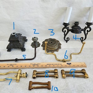 May include: A collection of vintage lamp parts, including ornate bases, decorative arms, and a double-light fixture. The components feature various finishes, such as bronze and brass, and are arranged on a white surface. A ruler is included for scale.