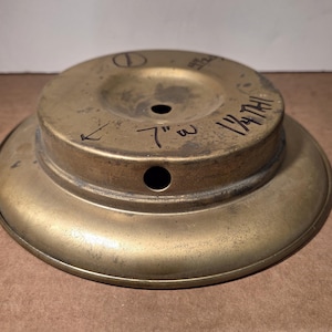 May include: A circular brass base with a stepped design, featuring a central hole and a side hole. Handwritten black ink markings on the surface indicate 7 inches wide and 1 1/4 inches high, with additional markings including a circle and a line.