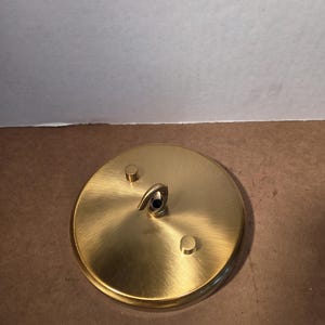 May include: A circular brushed brass ceiling canopy plate featuring a central hook and two small cylindrical knobs. A wooden ruler is positioned at the bottom, showing the plate has a diameter of approximately 12.7 cm across the metal surface.