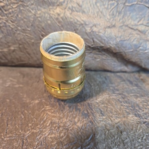 May include: A gold-colored lamp socket with a paper-lined interior and a silver-colored threaded base. The socket has a cylindrical shape with a ridged design around the base. The item is likely for a light fixture.