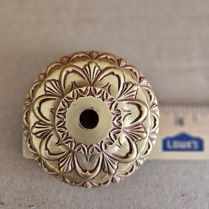 May include: A gold-coloured metal knob with a detailed floral design. The knob has a small hole in the centre.